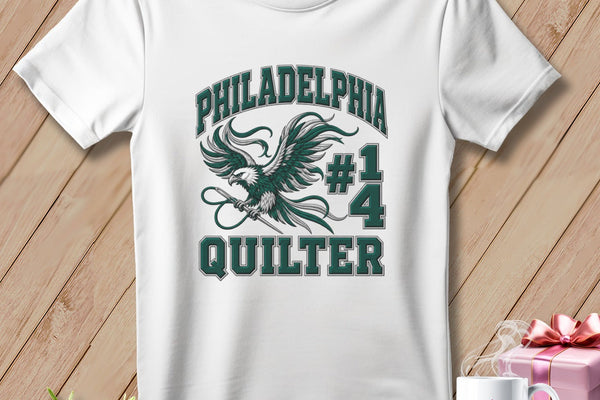 A white t-shirt hanging on a wooden background features a green eagle graphic and the text "Philadelphia #1 Quilter".