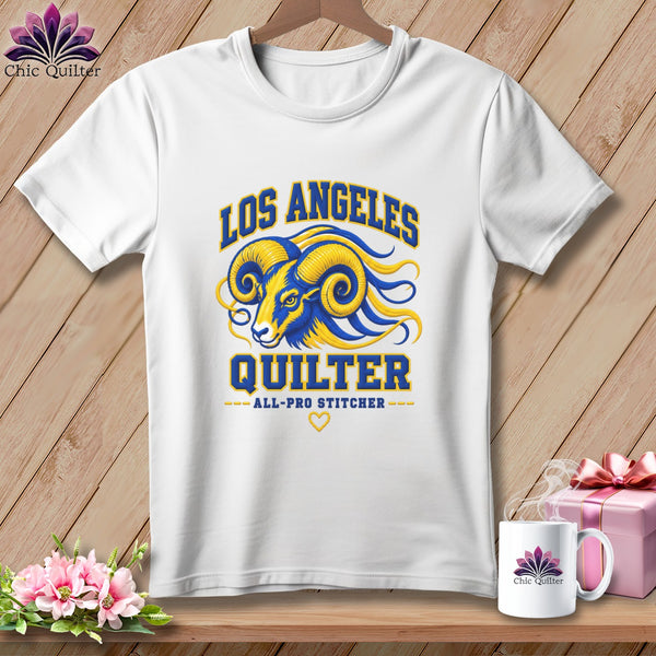 A white t-shirt hanging on a wooden background, featuring a blue and yellow ram graphic with text that reads Los Angeles Quilter.