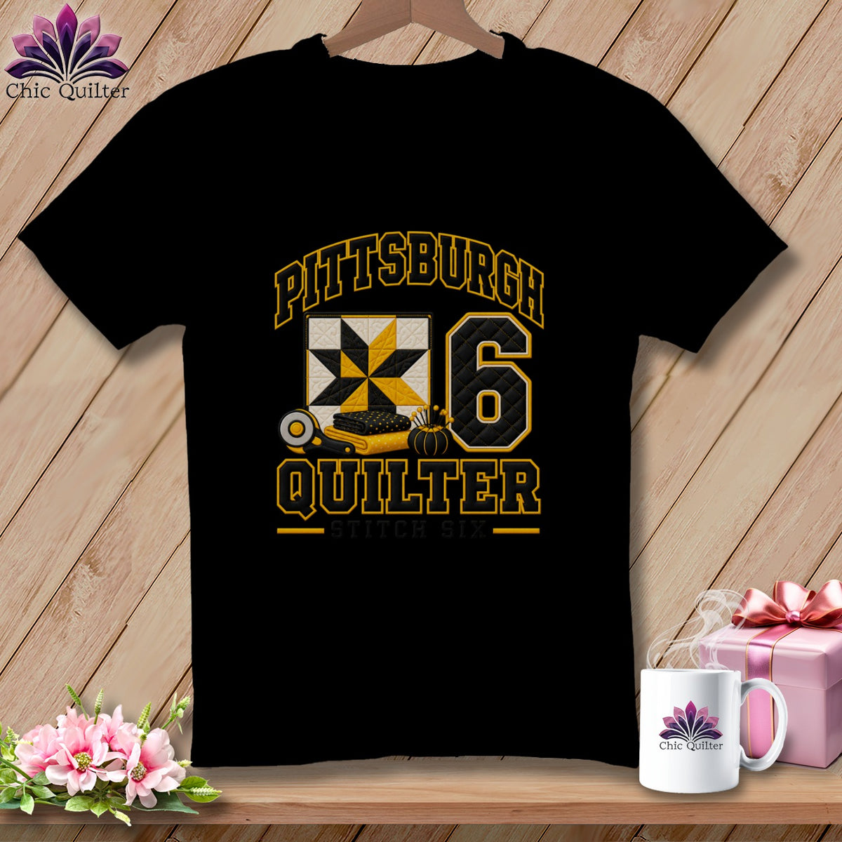 A black t-shirt hangs on a wooden hanger, featuring a yellow and black "Pittsburgh Quilter" graphic with a quilt block and the number six.