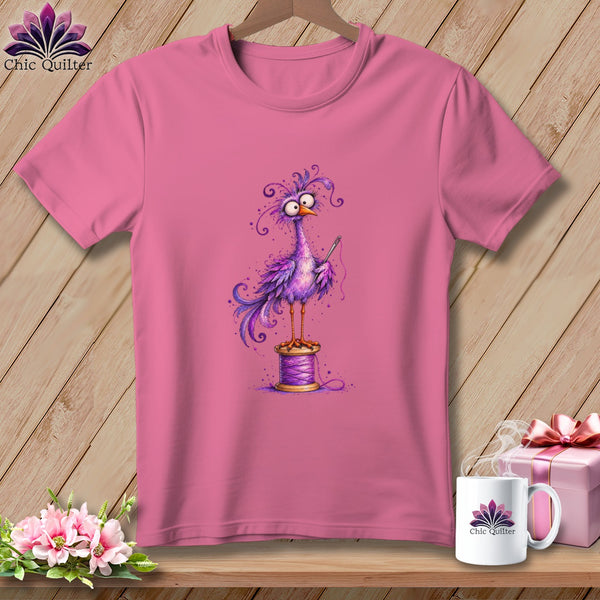 A pink t-shirt featuring a cartoon purple bird standing on a spool of thread while holding a needle.