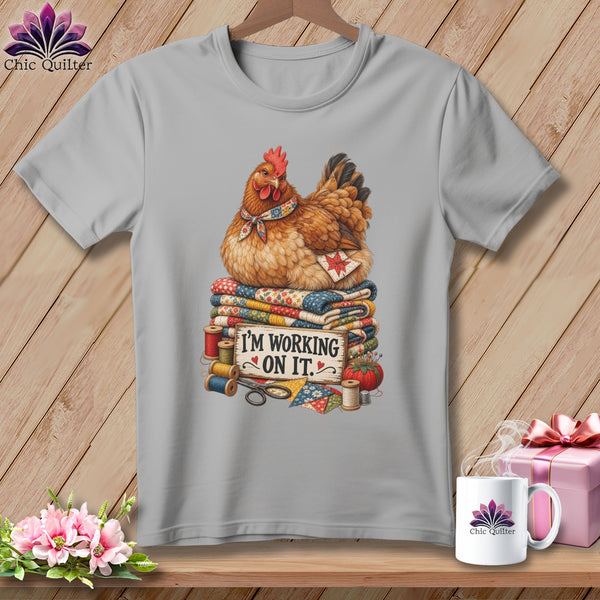 Chicken wearing a scarf sits on quilts with sewing supplies and text I'M WORKING ON IT.