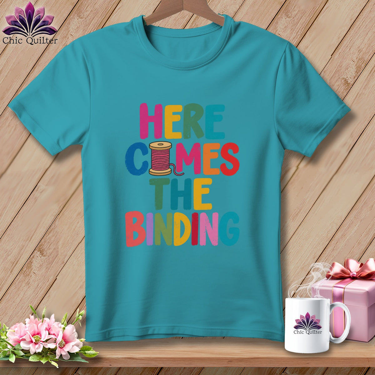 Turquoise t-shirt hanging on a wooden background, featuring the colorful text Here Comes the Binding and a spool of thread.