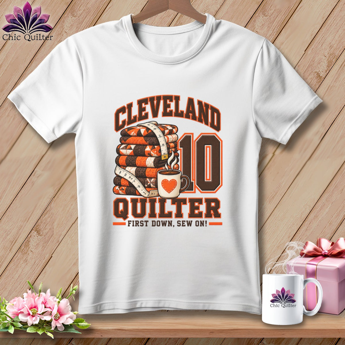 A white t-shirt features a graphic with a folded quilt, coffee mug, and text reading Cleveland Quilter First Down, Sew On.