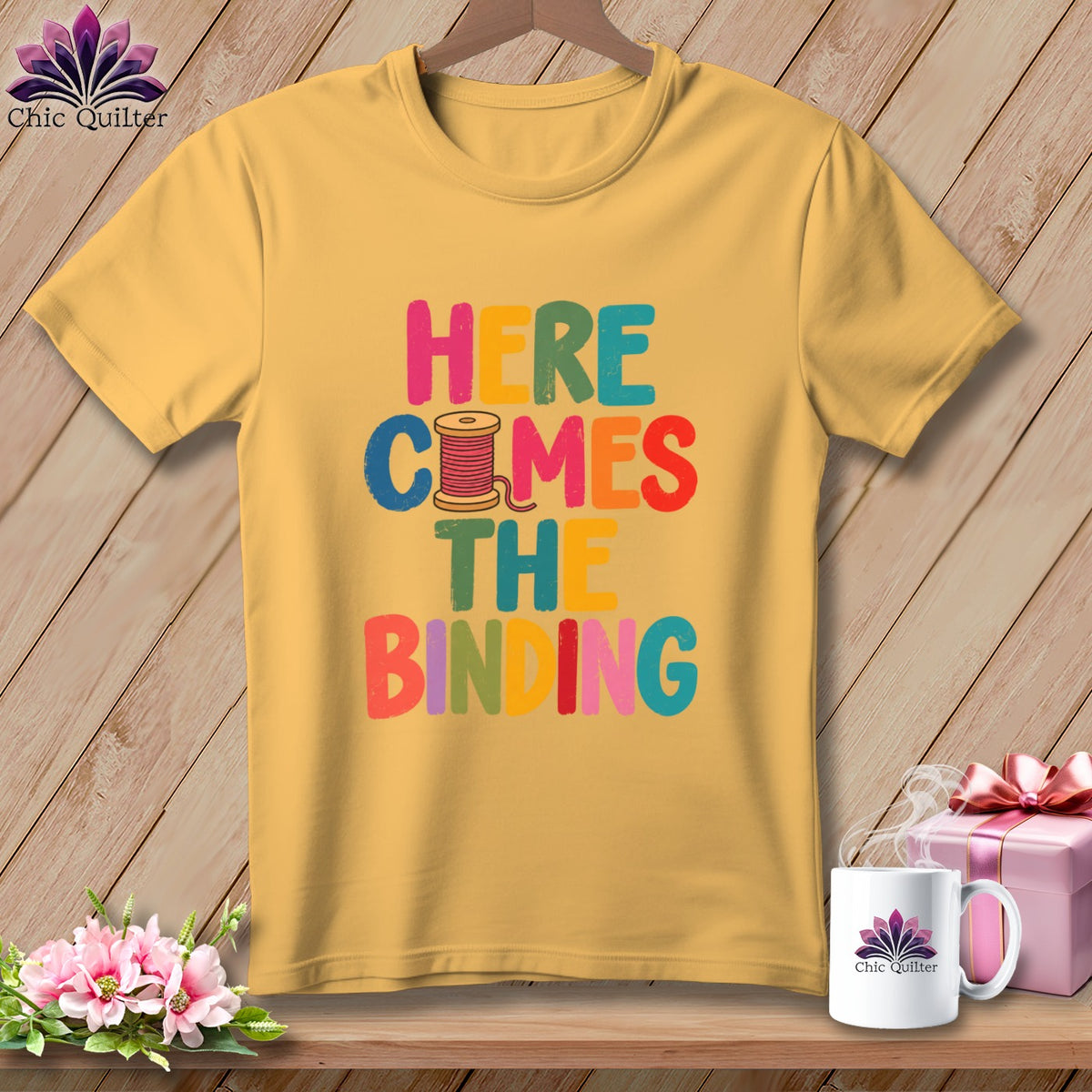 Yellow crewneck t-shirt featuring colorful text reading "HERE COMES THE BINDING" with a spool of thread.