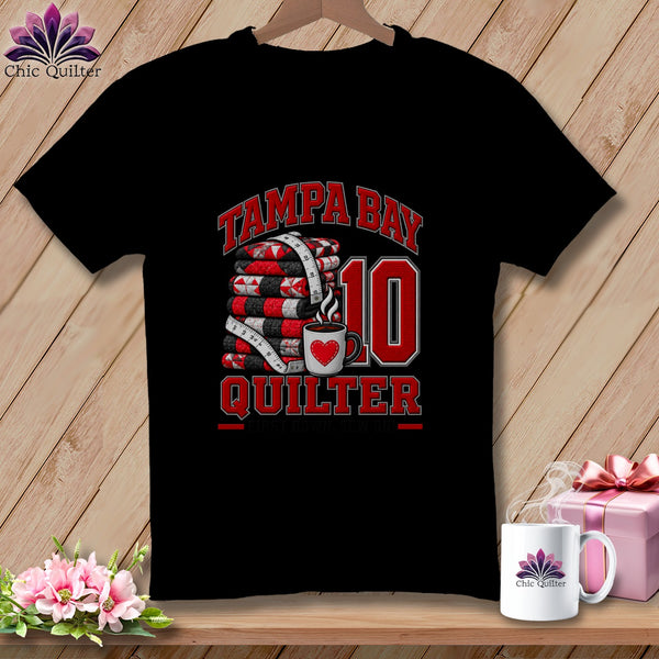 A black t-shirt features a graphic with a stack of quilts, a measuring tape, and a coffee mug with a red heart. Text reads Tampa Bay Quilter 10.