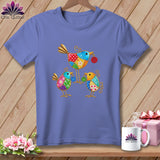 Periwinkle t-shirt on a wooden background, featuring a colorful folk art illustration of three birds with button accents.