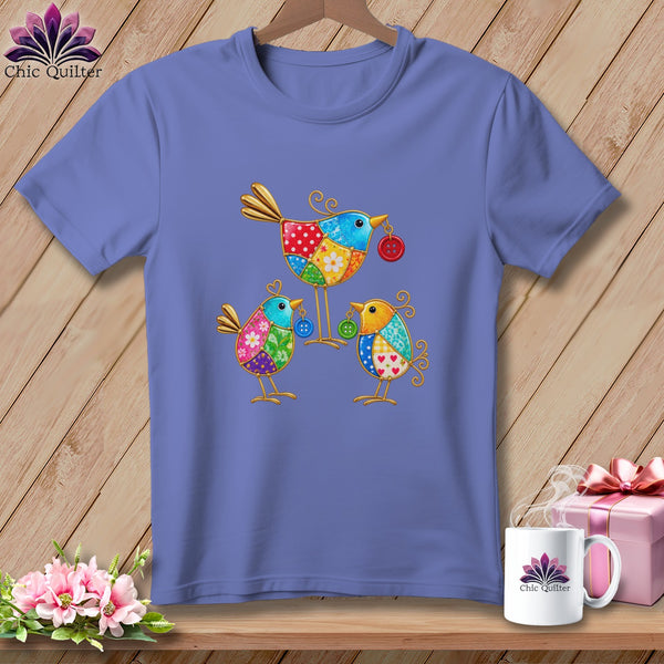 Periwinkle t-shirt on a wooden background, featuring a colorful folk art illustration of three birds with button accents.