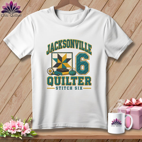 A white t-shirt hanging on a wooden background features a design with quilt-themed graphics and text.