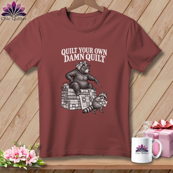 Maroon t-shirt featuring a black and white graphic of a bear and raccoon quilting, with text reading "Quilt Your Own Damn Quilt".