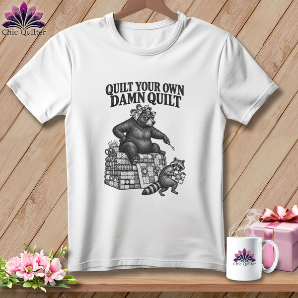 A white t-shirt featuring a black graphic of a bear and raccoon with a quilt and the text Quilt Your Own Damn Quilt.