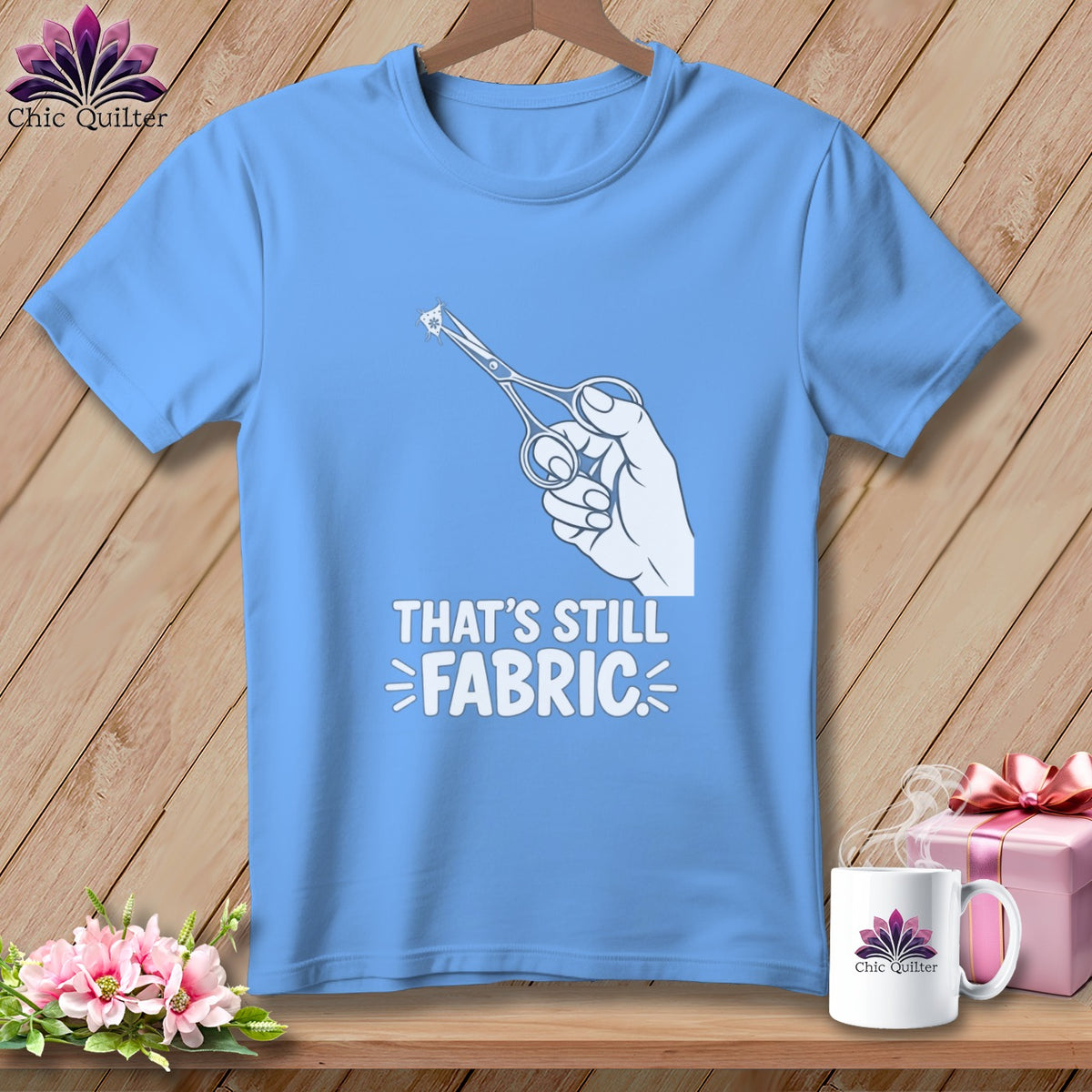 Blue t-shirt with white graphic of hand holding scissors cutting fabric, text reads "THAT'S STILL FABRIC".