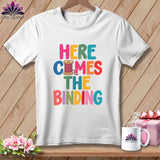 A white t-shirt hanging on a wooden background features colorful text reading Here Comes The Binding with a spool of thread graphic.