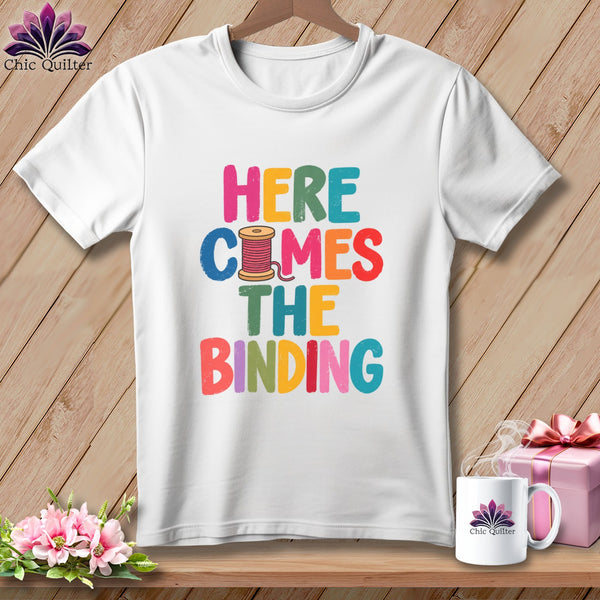 A white t-shirt hanging on a wooden background features colorful text reading Here Comes The Binding with a spool of thread graphic.