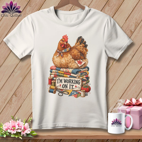 Chicken sitting on quilts with sewing supplies and "I'm working on it" text.