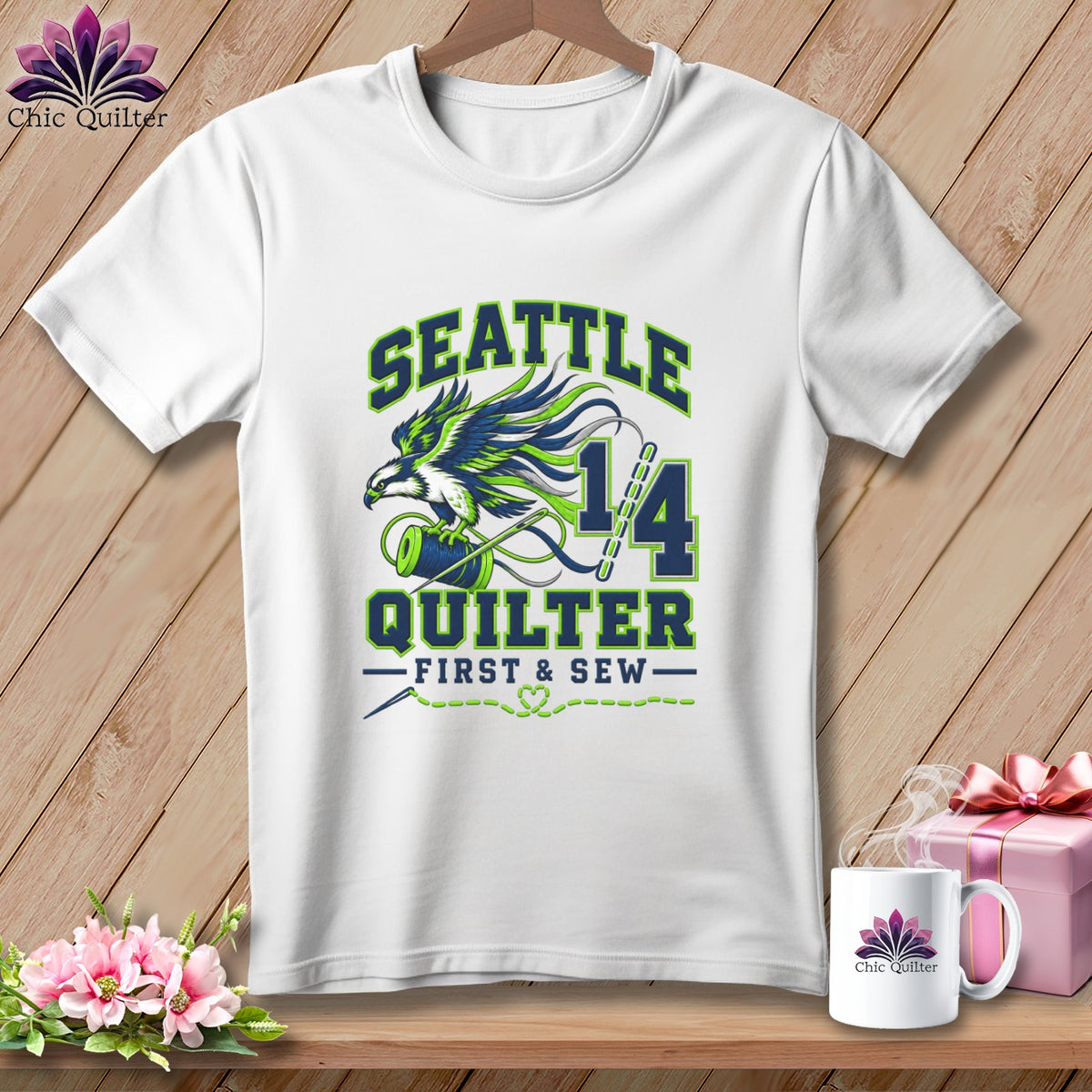 A white t-shirt hanging on a wooden background, featuring Seattle Quilter graphics with a bird and spool.