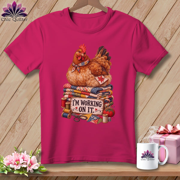 Chicken sitting on quilts with sewing notions and text "I'M WORKING ON IT."