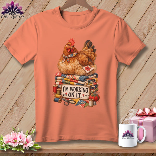 Salmon t-shirt with a chicken sitting on quilts and the text "I'M WORKING ON IT."