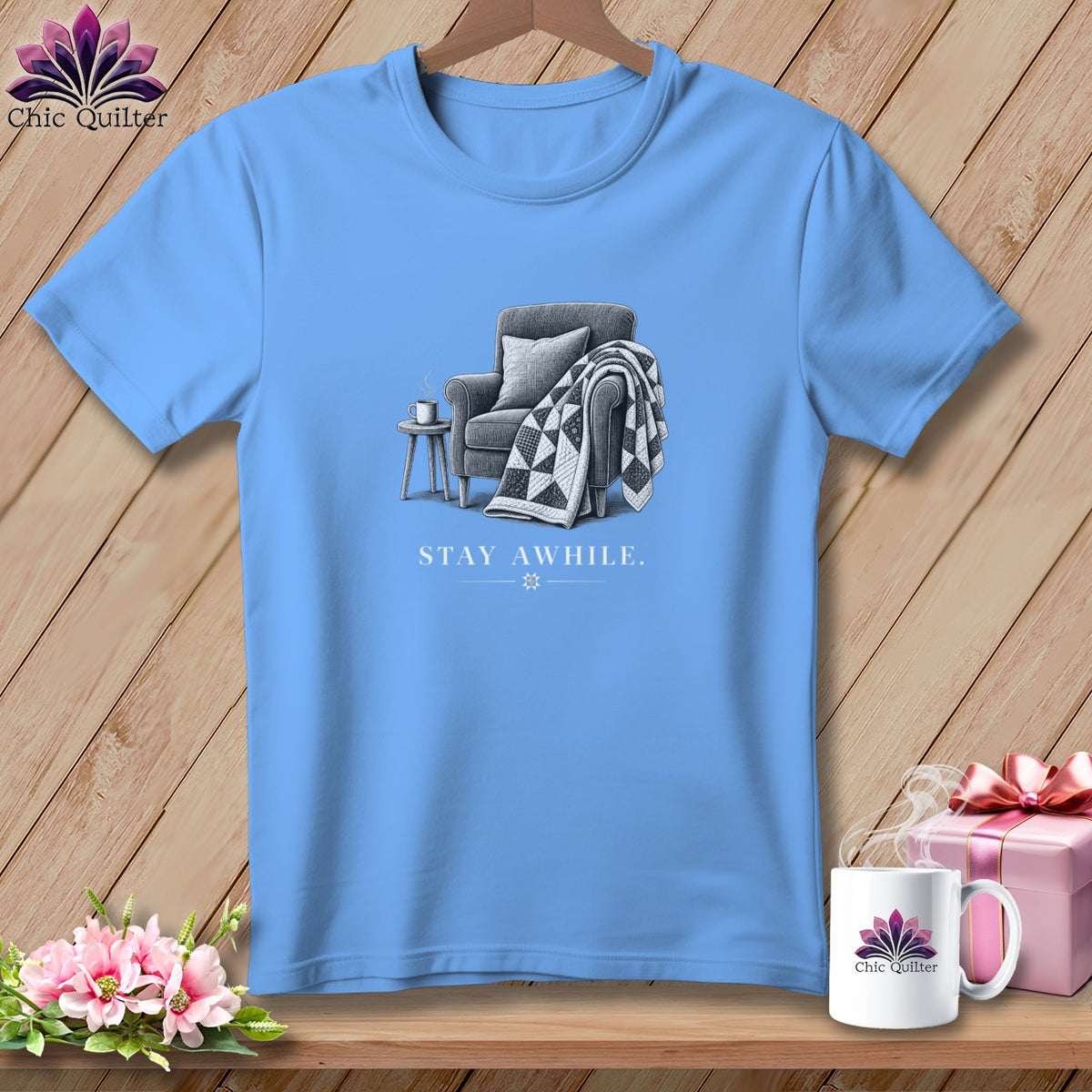 Light blue t-shirt with a drawing of a chair, quilt, and mug, reading "Stay Awhile".
