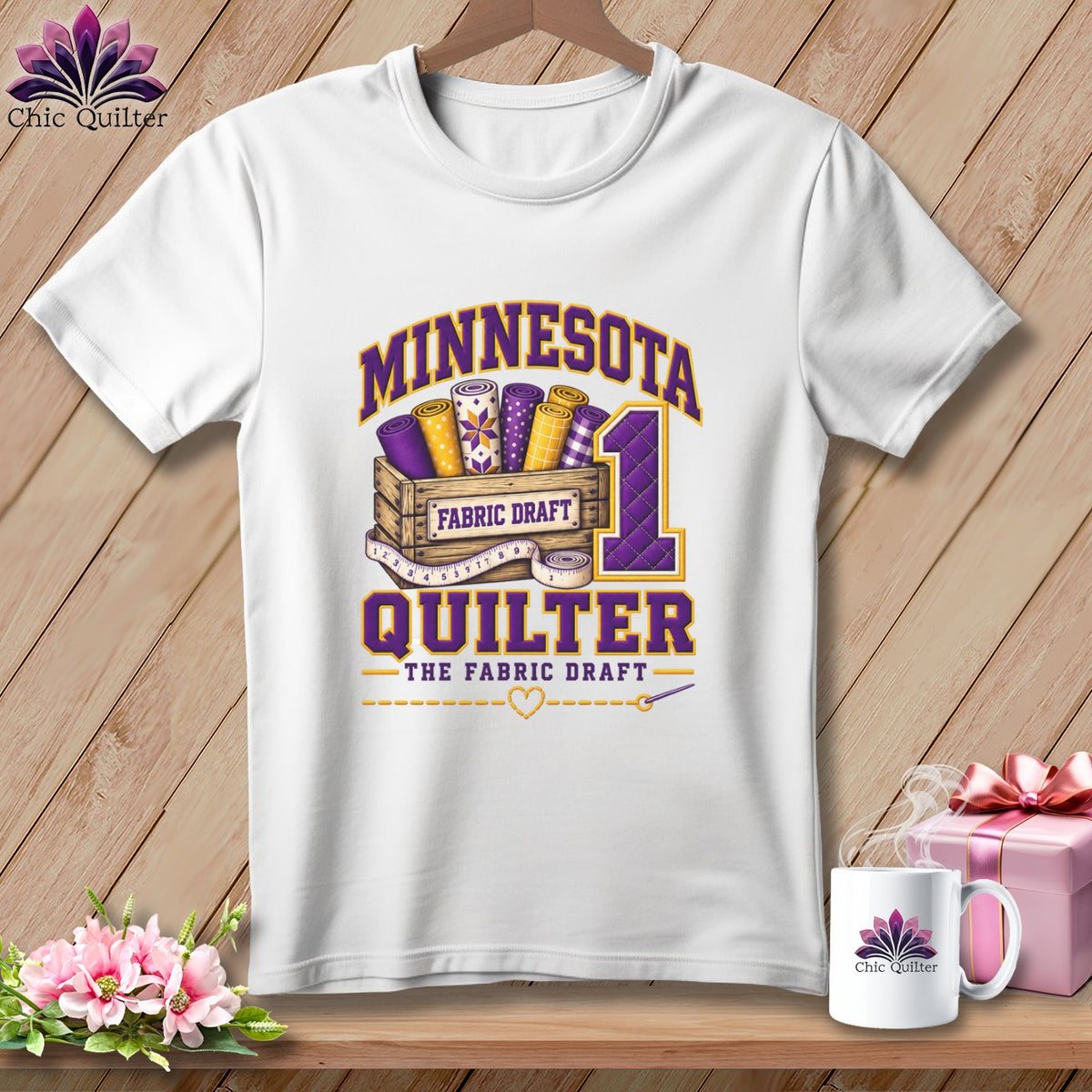 A white t-shirt hanging on a wooden wall features a graphic with the text "MINNESOTA QUILTER THE FABRIC DRAFT".