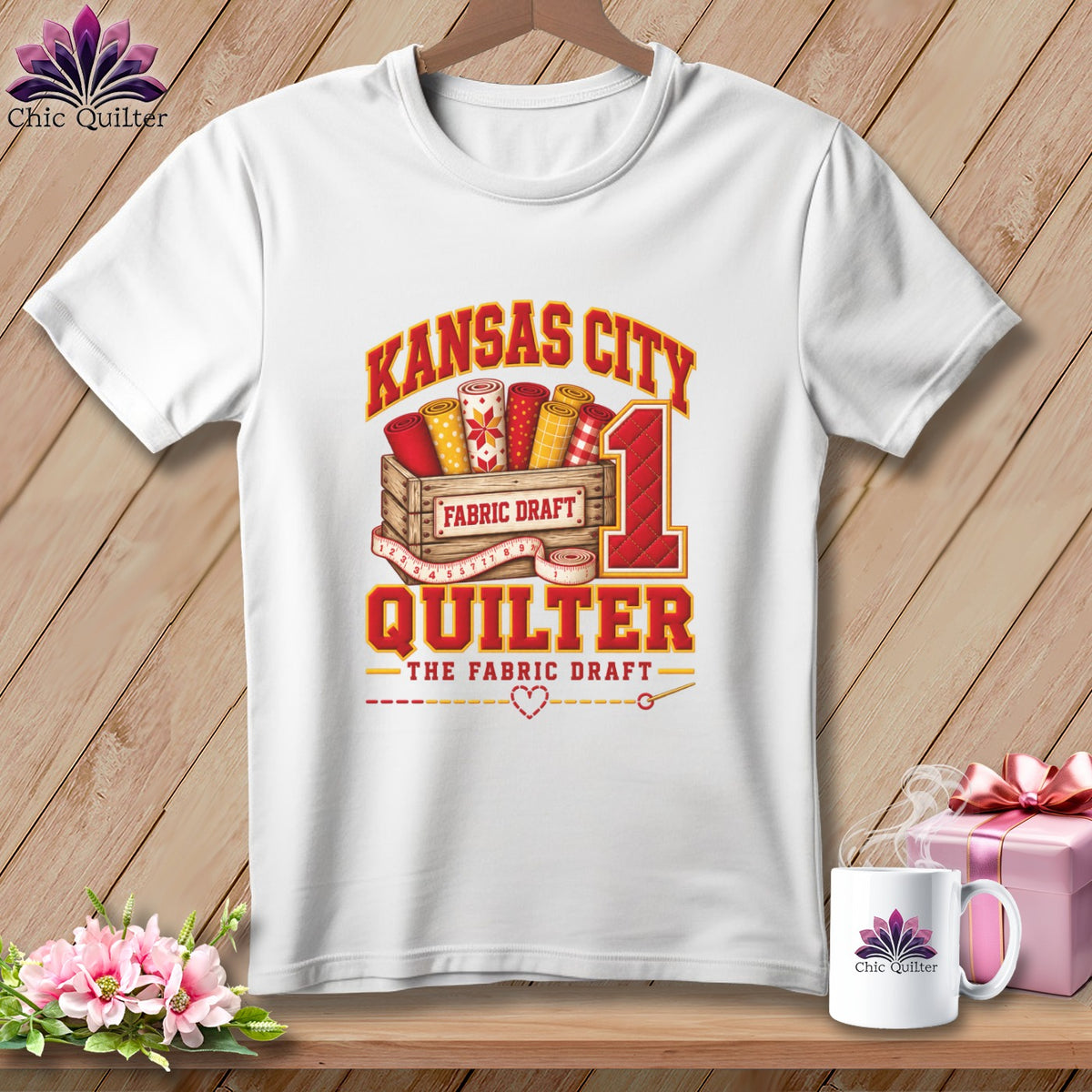 A white short-sleeved t-shirt featuring a Kansas City Quilter graphic with fabric rolls in a wooden crate.