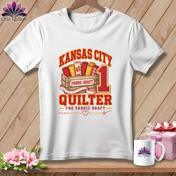 A white short-sleeved t-shirt featuring a Kansas City Quilter graphic with fabric rolls in a wooden crate.