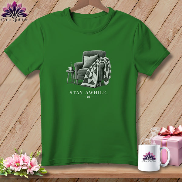 Green t-shirt with an illustration of a chair, blanket, table, and coffee mug. Text reads "STAY AWHILE.".