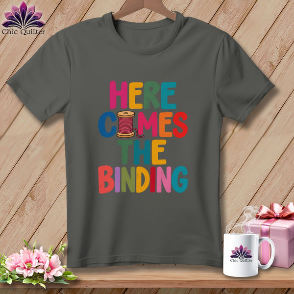 Dark gray t-shirt with multicolored text reading Here Comes The Binding and a thread spool graphic.