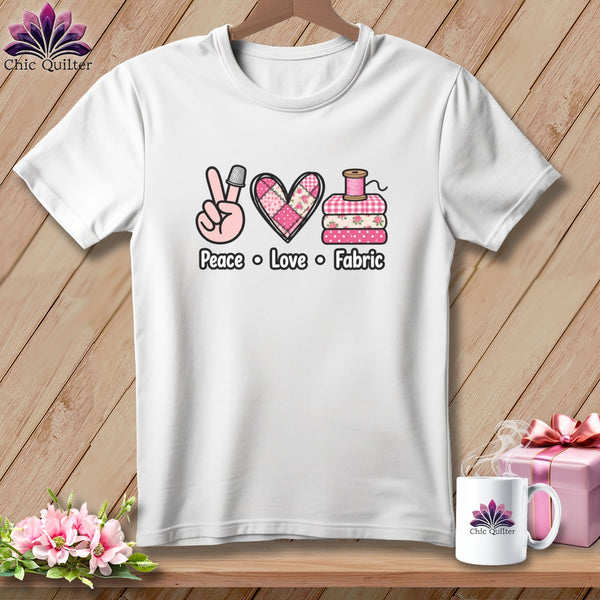 A white t-shirt hanging on a wooden background, featuring a pink graphic with the text Peace Love Fabric.