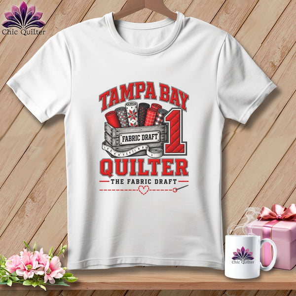 White t-shirt featuring a red and black Tampa Bay Quilter graphic with fabric rolls.