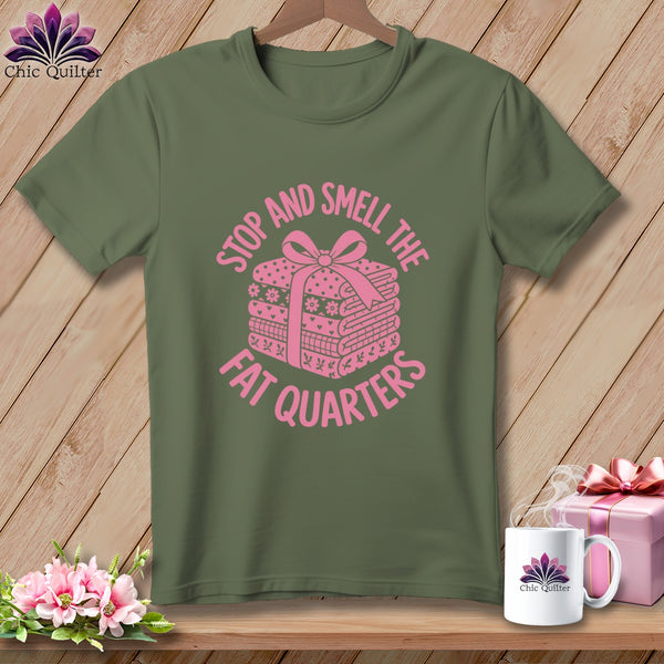 An olive green t-shirt with pink text reading STOP AND SMELL THE FAT QUARTERS surrounding a stack of folded fabric tied with a bow.