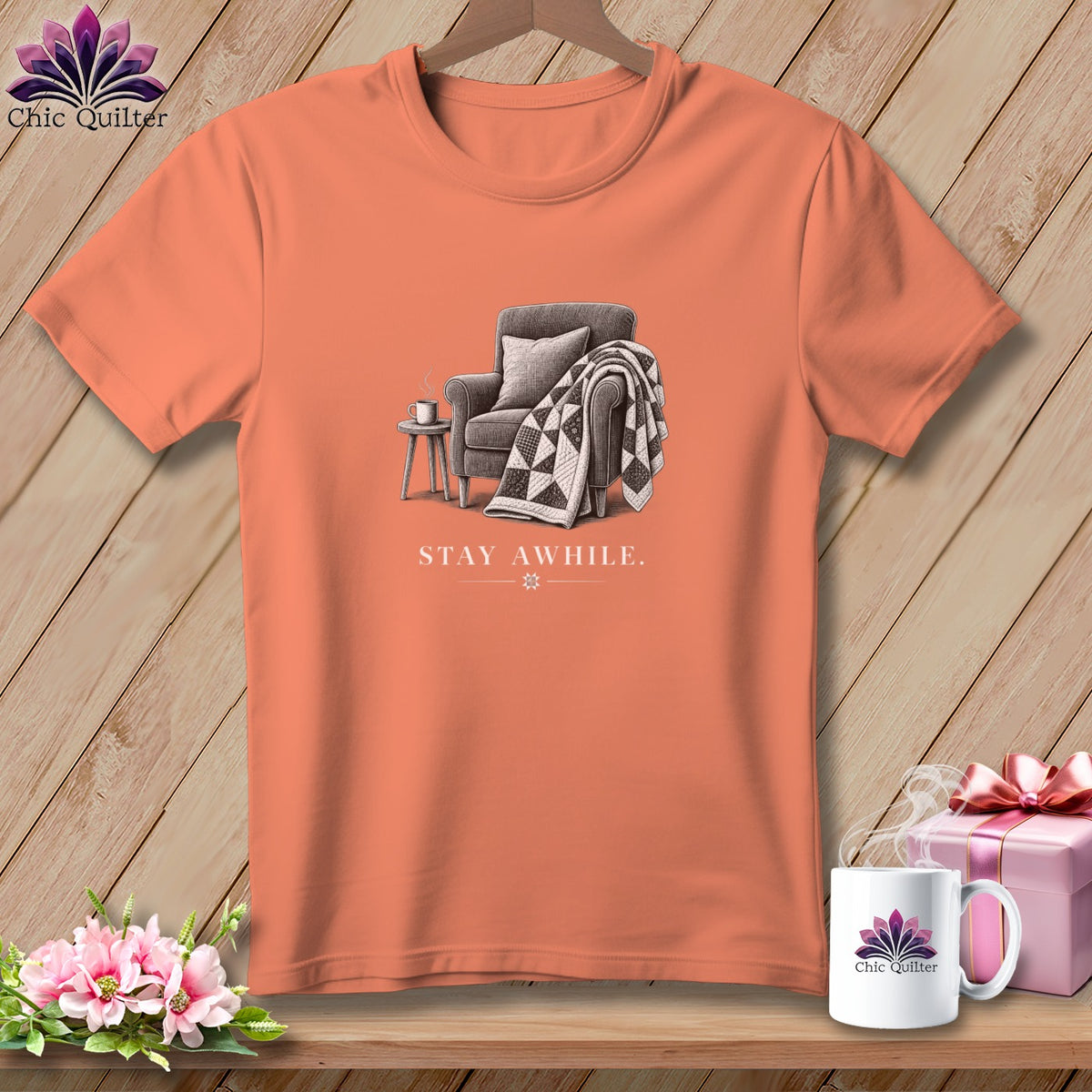 Peach t-shirt with an illustration of a chair, quilt, and mug with "STAY AWHILE" text.