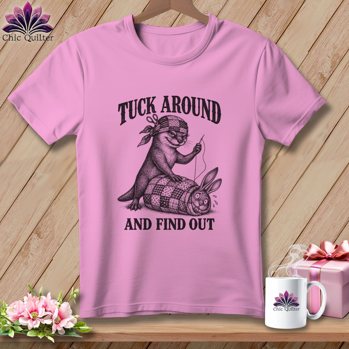 Pink t-shirt with a black graphic of an otter sewing a rabbit into a quilt, featuring the text Tuck Around And Find Out.