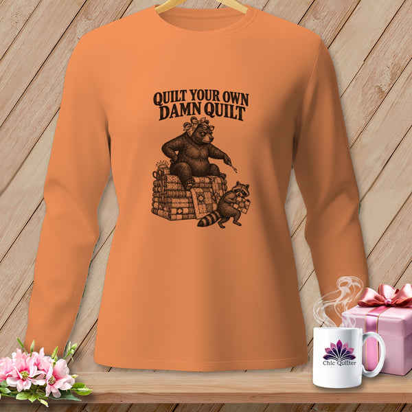 An orange long-sleeve shirt with a graphic of a bear and raccoon quilting and the text Quilt Your Own Damn Quilt.