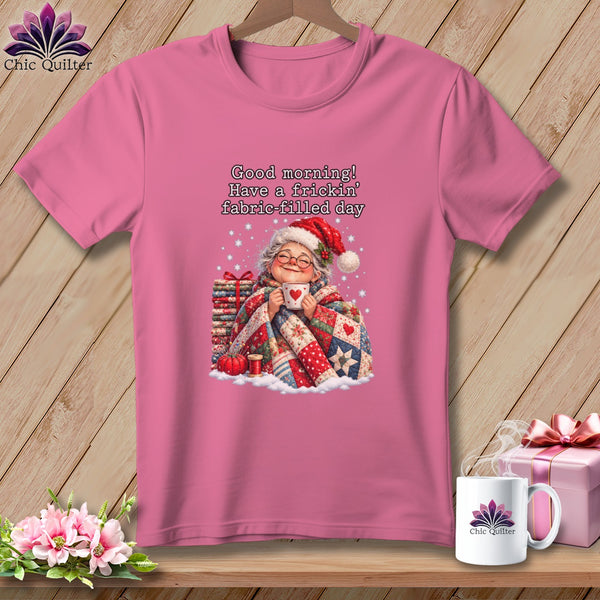 A pink crewneck t-shirt featuring an illustration of an elderly woman holding a mug, wrapped in a quilt, beneath the text "Good morning! Have a frickin' fabric-filled day."