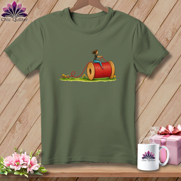 An olive green t-shirt featuring an illustration of a woman riding a large spool of red thread.