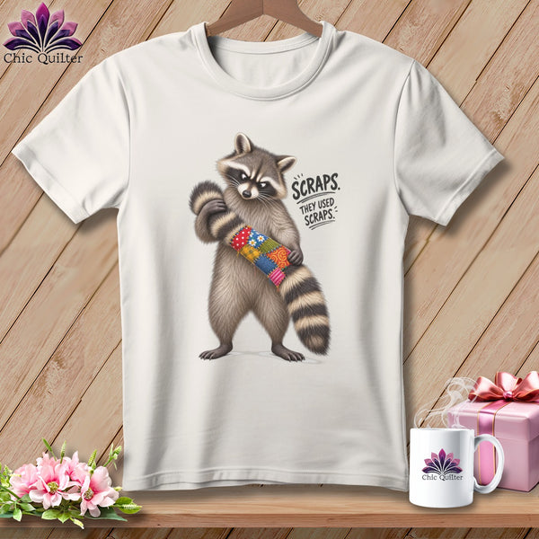 White t-shirt with raccoon holding colorful quilt scraps, text "SCRAPS THEY USED SCRAPS."