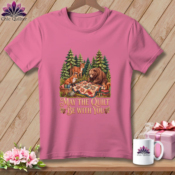 A pink crewneck t-shirt featuring forest animals gathered around a colorful patchwork quilt with the text May the Quilt Be with You.