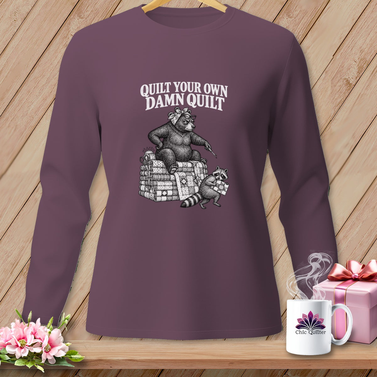 A dark purple long-sleeved shirt featuring a vintage-style graphic of a bear and raccoon quilting, with text that reads QUILT YOUR OWN DAMN QUILT.