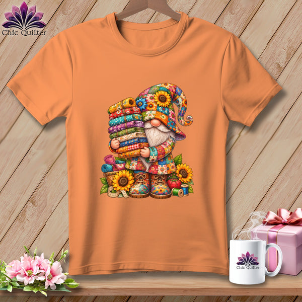 Orange t-shirt featuring a colorful gnome graphic holding a tall stack of folded fabrics, surrounded by sunflowers.