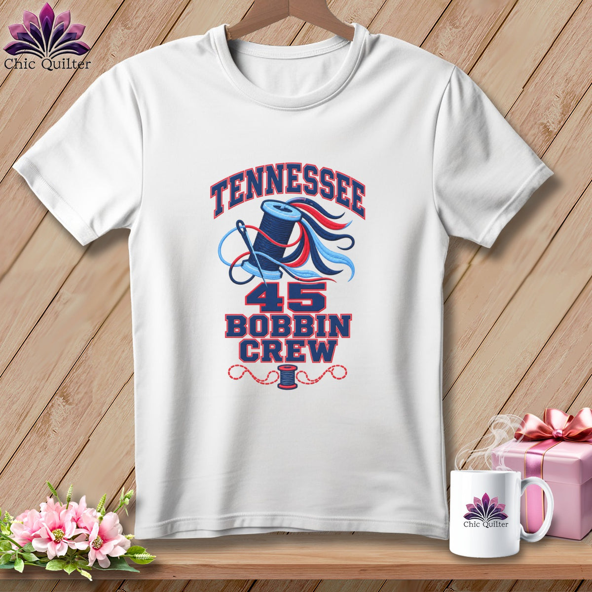 A white t-shirt hanging on a wooden background features a graphic with the text Tennessee 45 Bobbin Crew, a spool of thread, and a needle.