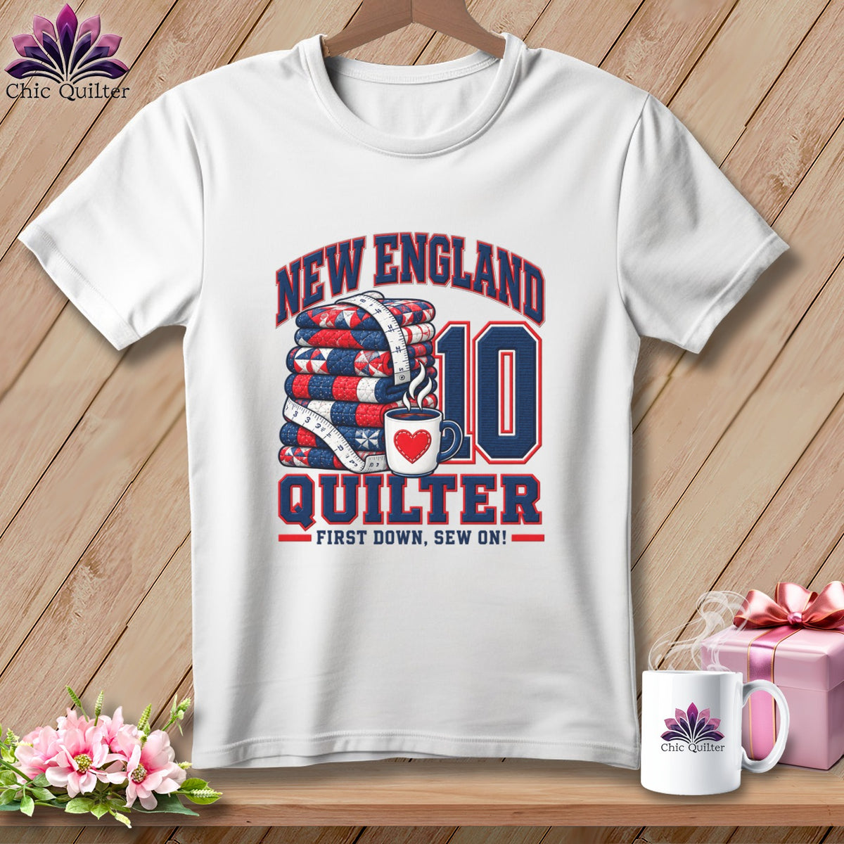 A white t-shirt hanging on a wooden background features a New England Quilter graphic with a stack of quilts, a measuring tape, a coffee mug, and the number 10.