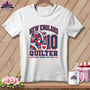 A white t-shirt hanging on a wooden background features a New England Quilter graphic with a stack of quilts, a measuring tape, a coffee mug, and the number 10.