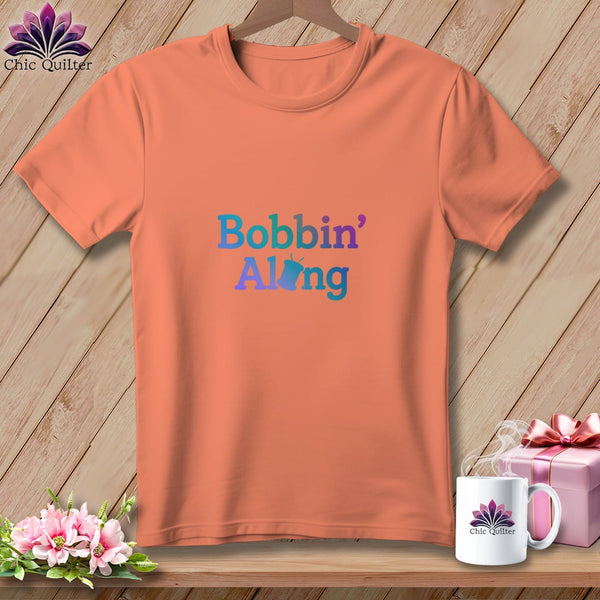 Peach t-shirt with gradient text reading "Bobbin' Along" and a small bobbin graphic.