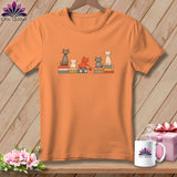 Orange t-shirt featuring five illustrated cats sitting on colorful patterned blocks against a wooden background.