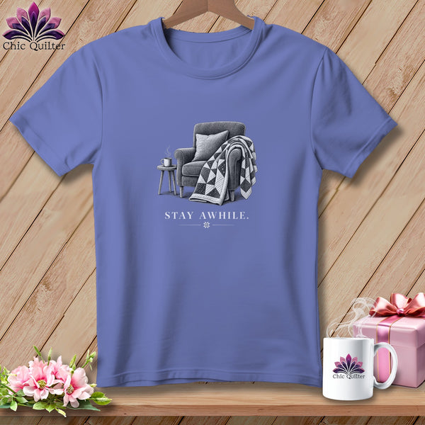 Purple t-shirt with a graphic of a chair, quilt, and mug, reading "STAY AWHILE."