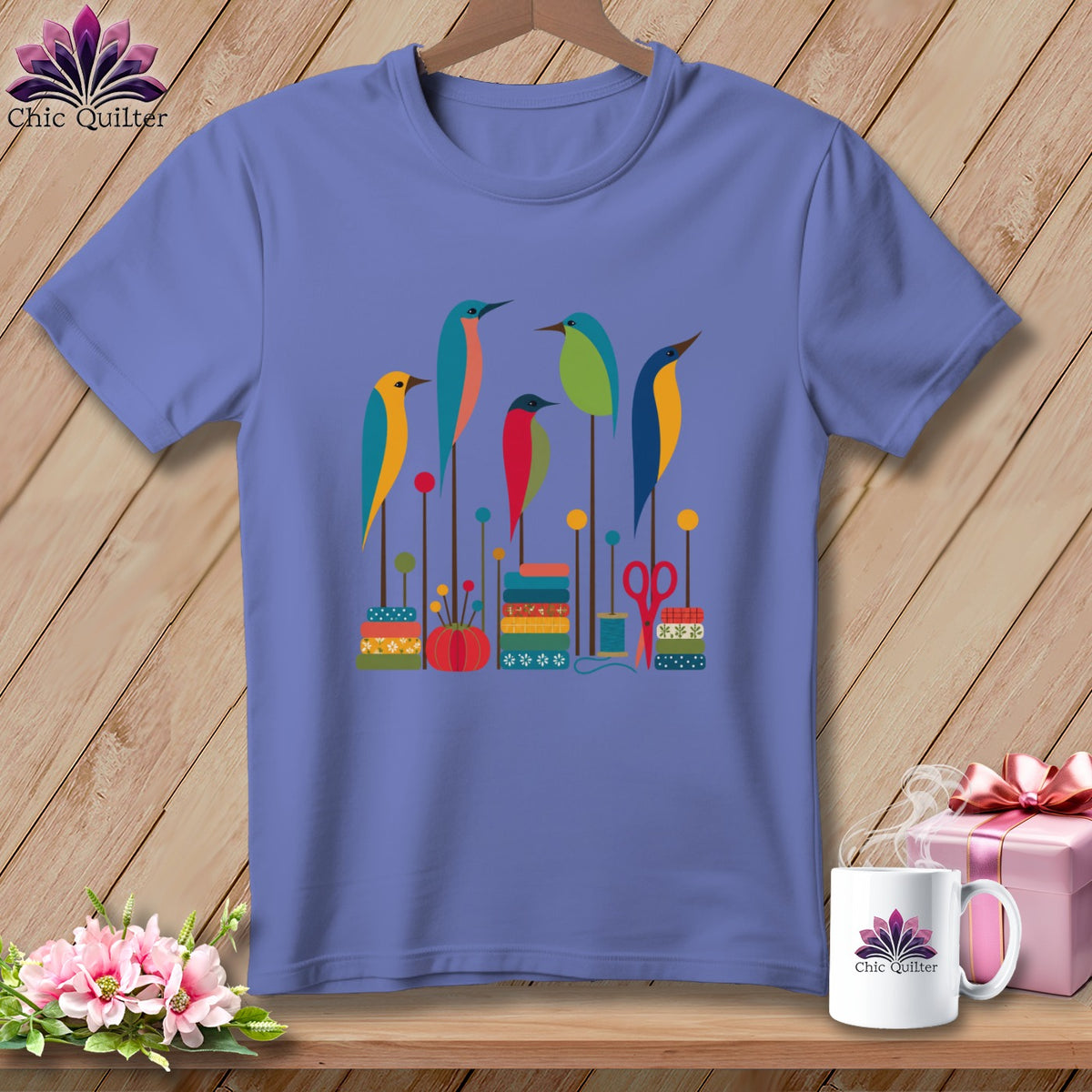 Periwinkle short-sleeve t-shirt with a graphic of colorful stylized birds, stacked spools of thread, and sewing accessories.