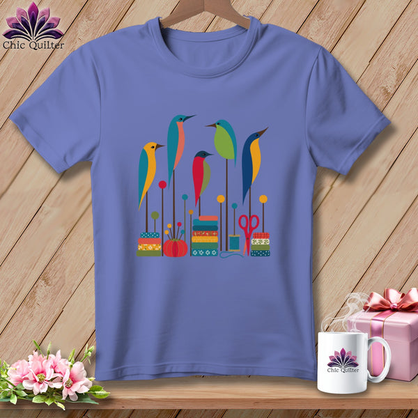 Periwinkle short-sleeve t-shirt with a graphic of colorful stylized birds, stacked spools of thread, and sewing accessories.