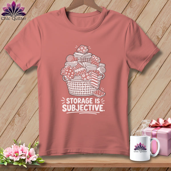 Pink t-shirt with a graphic of a basket overflowing with fabric rolls and stacks, text reads "STORAGE IS SUBJECTIVE."