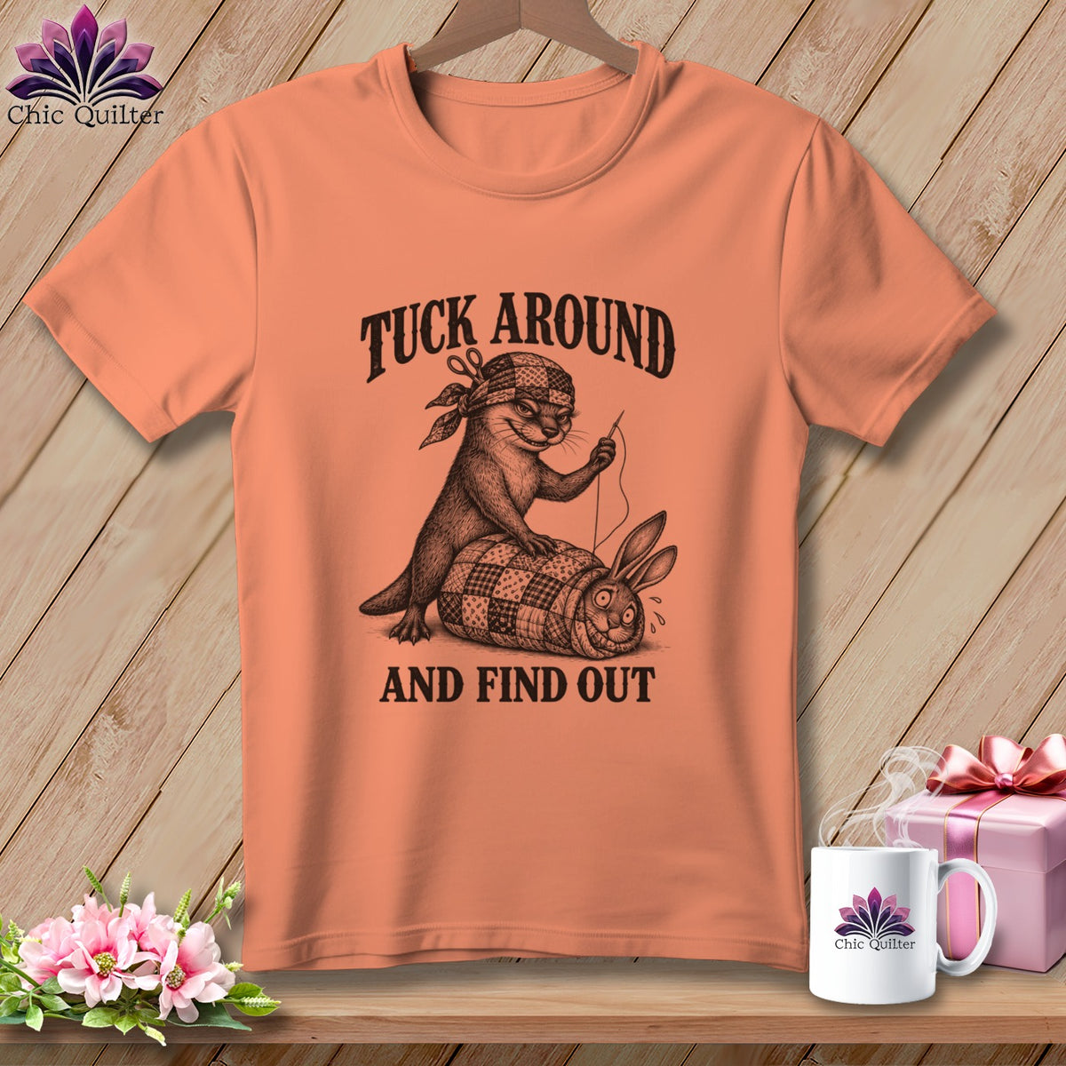 Peach t-shirt with a humorous graphic of an otter sewing a rabbit into a quilt.