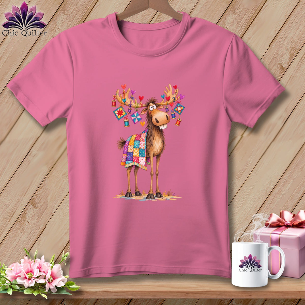 Pink t-shirt featuring a cartoon moose wearing a patchwork blanket, decorated with hearts and quilting supplies.